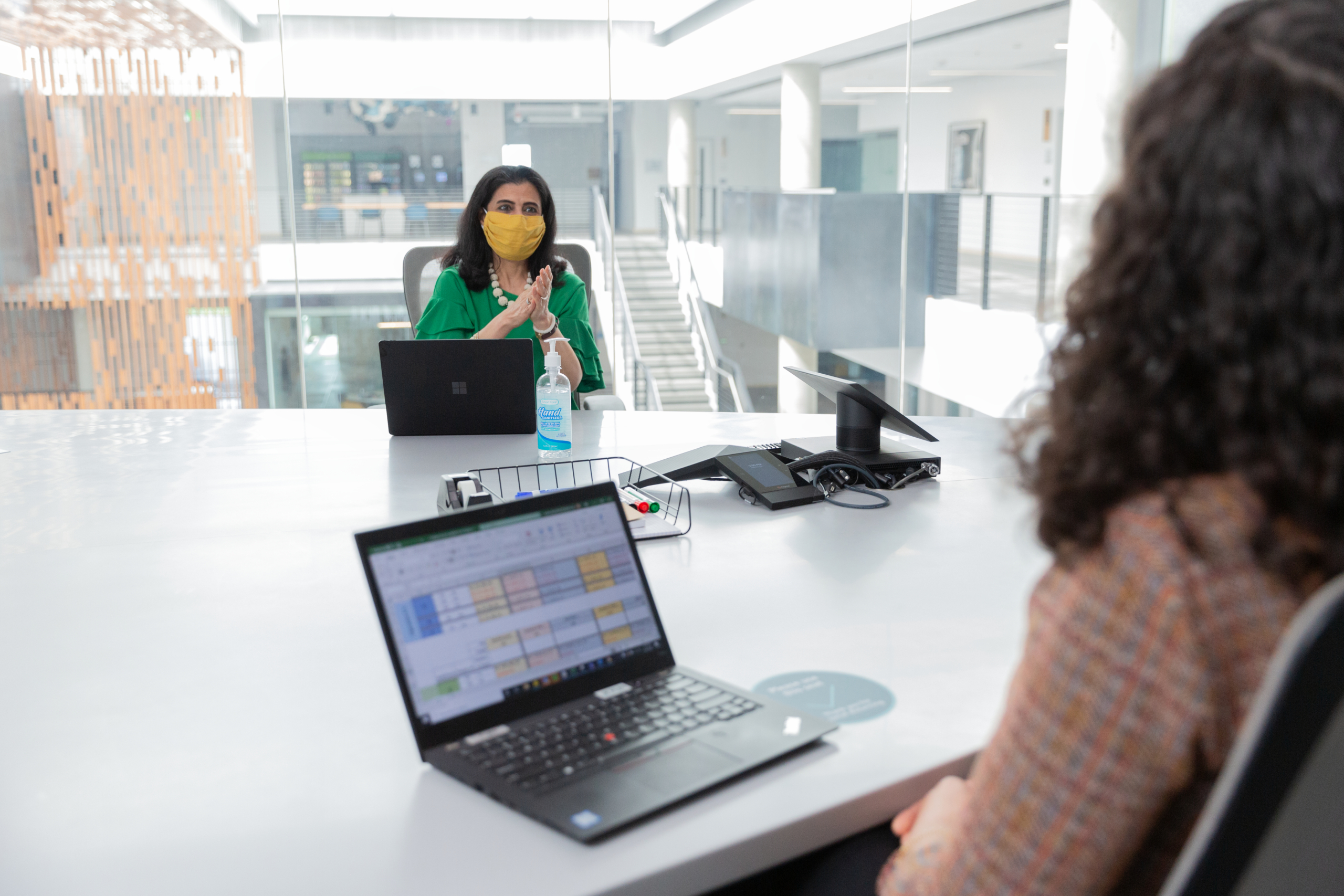Two female employees wearing face masks and talking in a conference room while social distancing. Hybrid Workplace collection. Keywords: Hybrid Workplace, Hybrid Worksite, Return to Workplace, Return to Work, Return to Worksite, Return to Office, Return to Campus, Reopening, Back to work, COVID-19, COVID-19 policies, COVID, Coronavirus, Pandemic, Health, Health and Safety, Face Covering, Face Mask, Masks, Masks at Work, Masks in Workplace, Open Workspace, Open Workspace Policies, Social Distancing, Social Distancing Policies, Social Distance, Physical Distancing