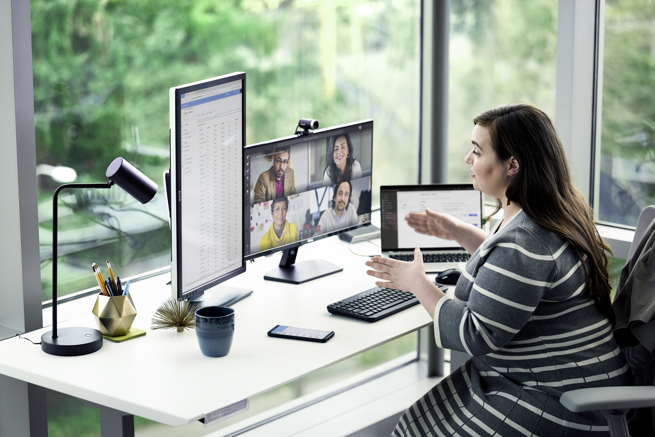 Female enterprise employee working at desk with multiple devices, including HP Elitebook, running a Microsoft Teams conference call. Two different screen images are available: one featuring 4 people on screen, another with 9 people on screen