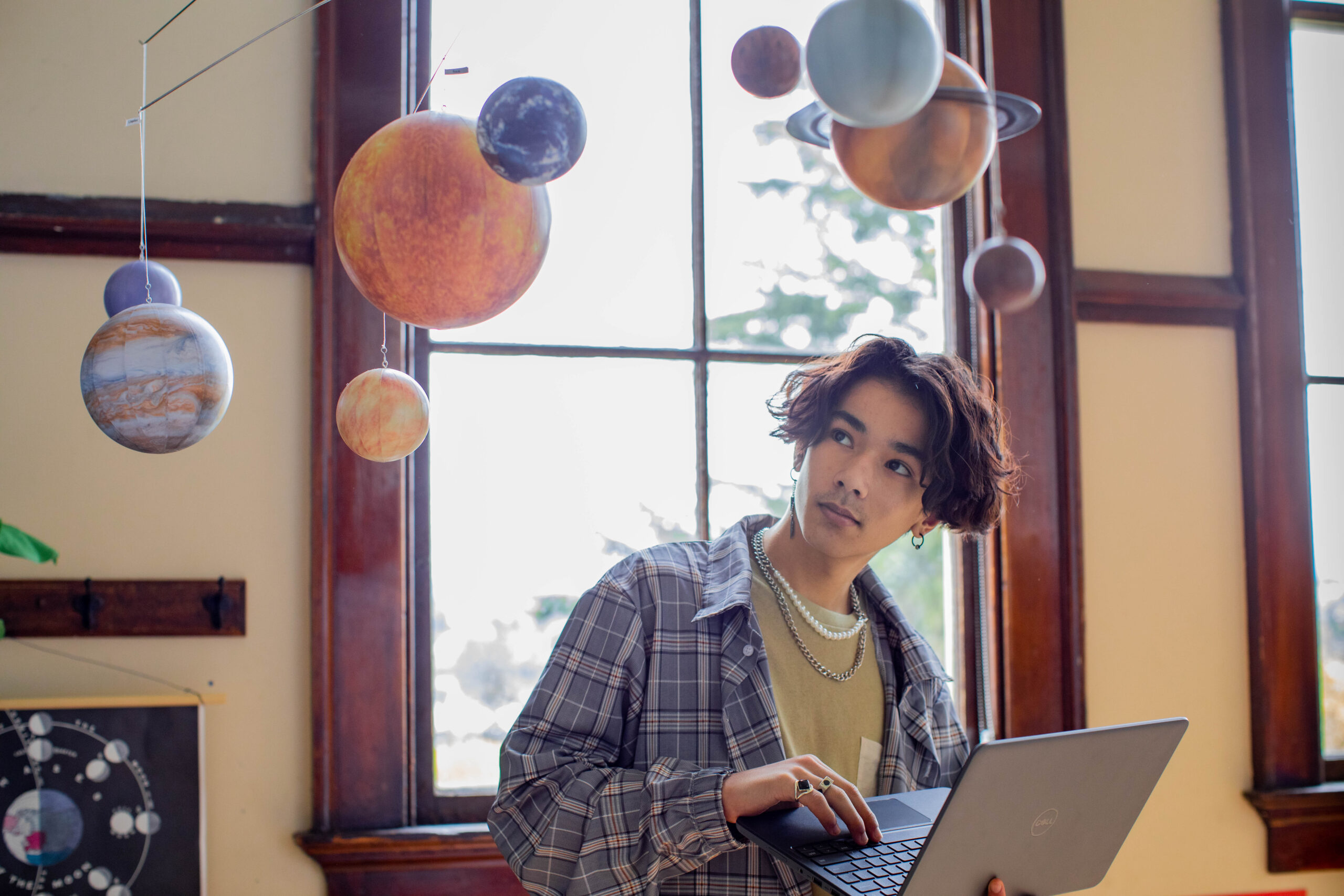 Photo of a student in a classroom working on an open laptop and looking at a solar system mobile hanging from the ceiling