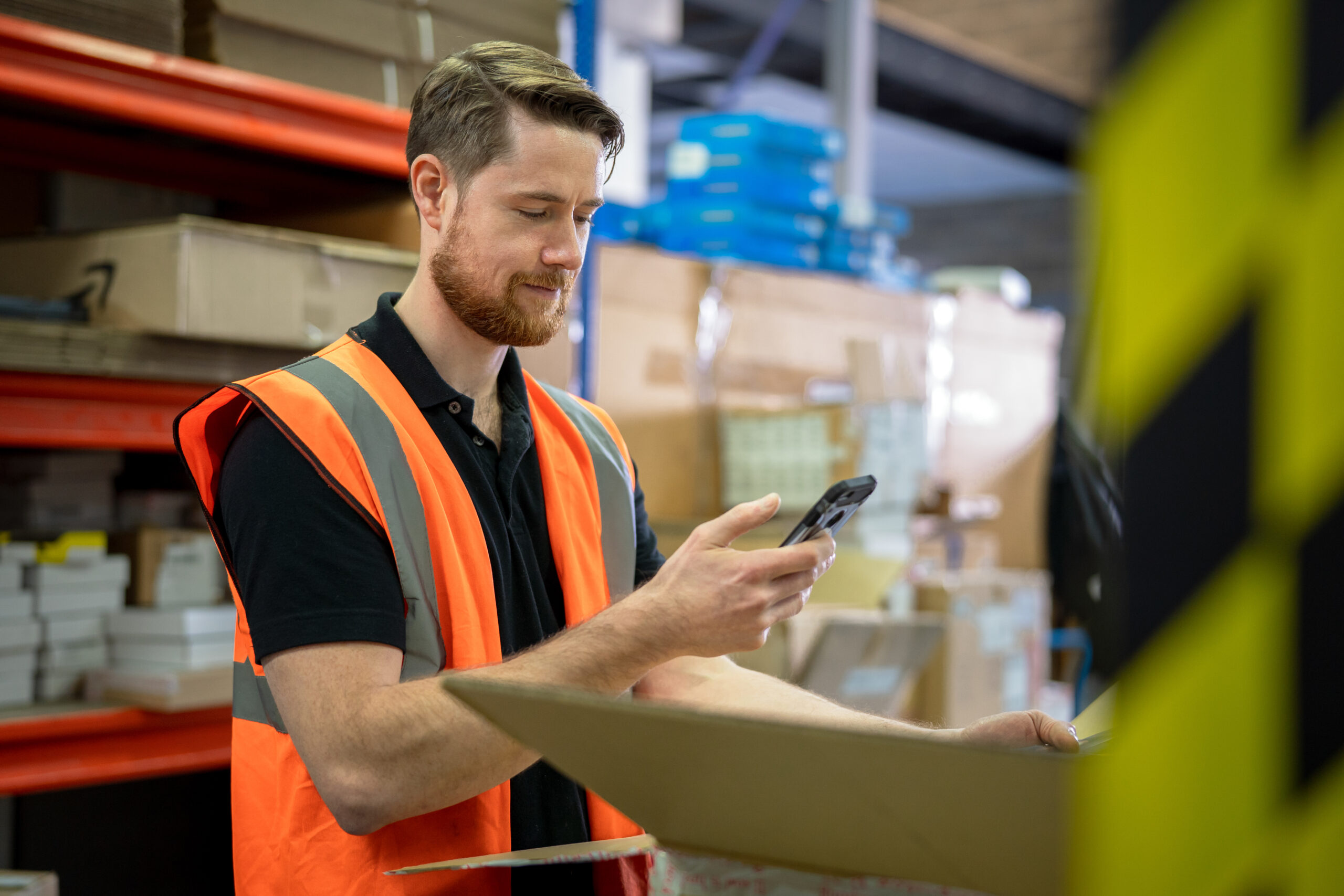Photo of a man wearing an orange vest in a warehouse who is doing inventory on his mobile phone