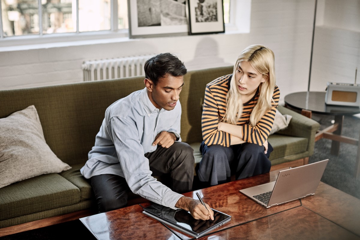 A man and women sitting on a couch in an office setting, collaborating on a project with their Lenovo Yoga; one set up as a Laptop and the other being used as a tablet