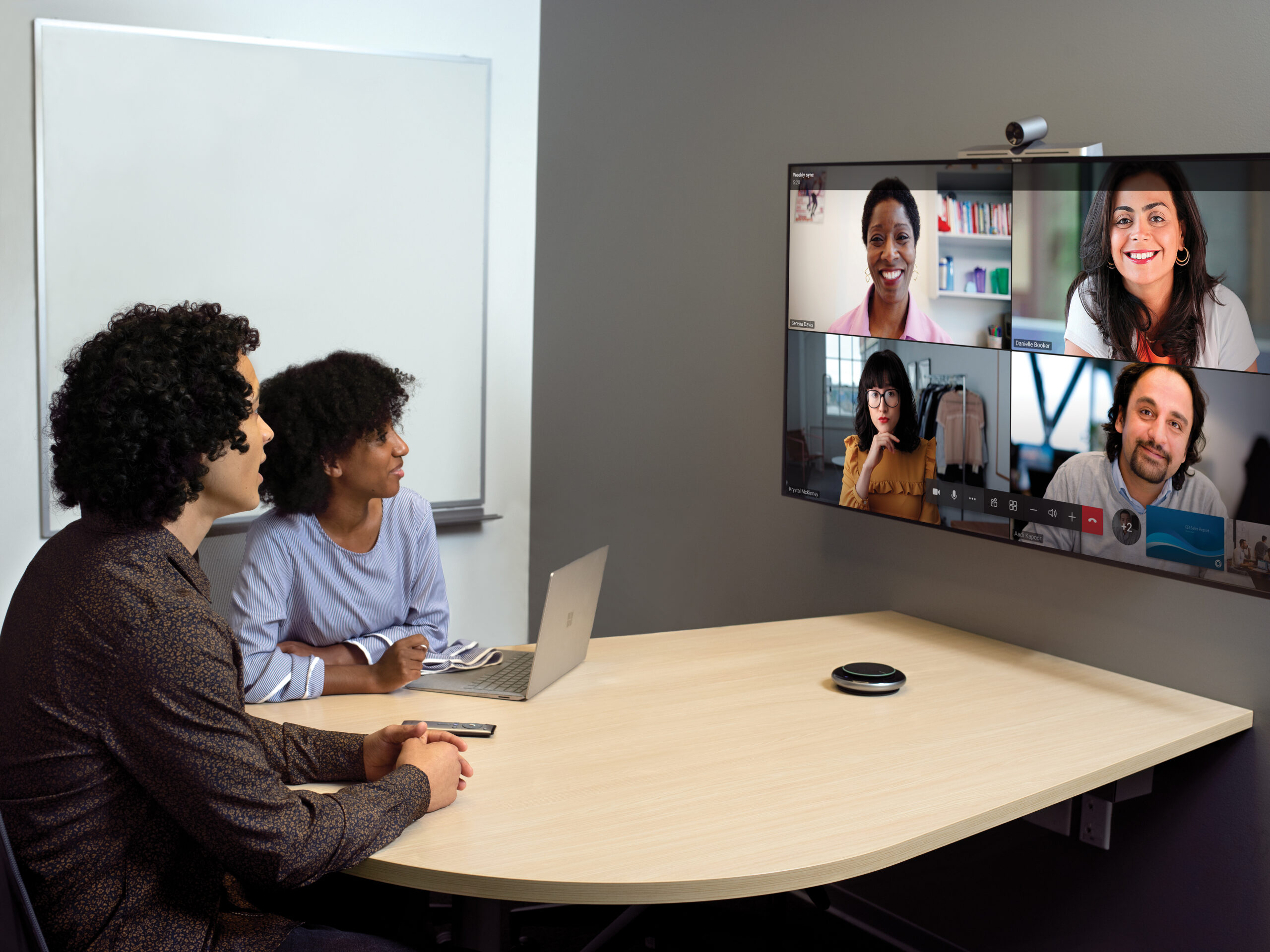 One male and one female in small conference room with a Yealink Microsoft Teams Room on Android device being used for remote video meeting with Teams Meetings. Large mounted display showing remote participants and one Surface Device in view