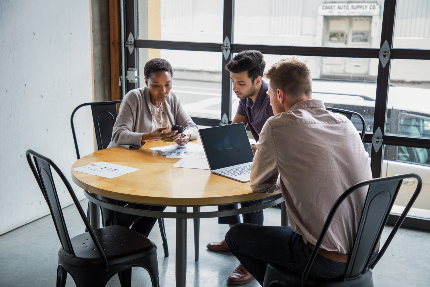 One female and two male workers sitting at round table in casual office setting or café/coffeehouse. The woman is holding a phone to show something to one of the men. The other man is using a Surface Laptop