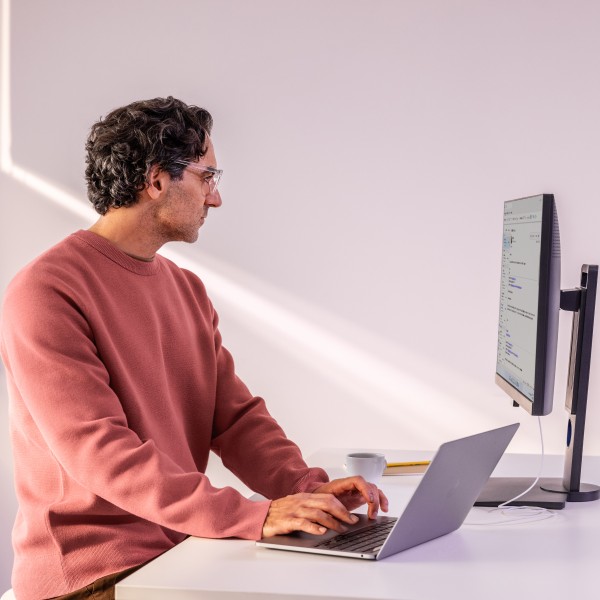Man in focused work, standing at a desk working on a laptop and looking at a monitor