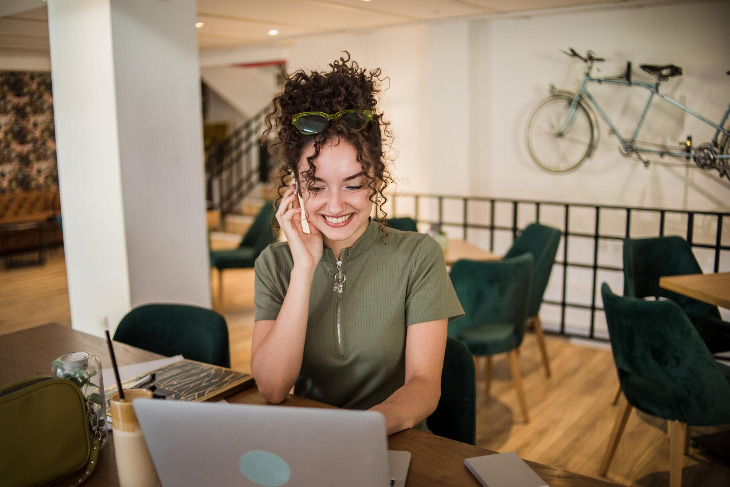 A young woman is working on a laptop while sitting in a cafe and drinking a frappe coffee that is on the table