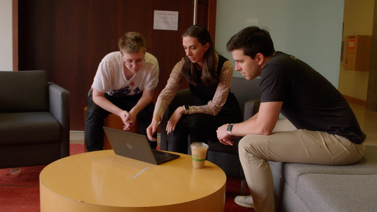 An IT professional and two students collaborating on a laptop outside of an office at a university