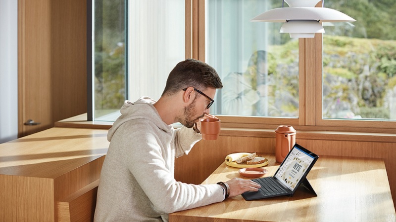 Ein Mann sitzt mit einem Laptop am Tisch, er trinkt dabei aus einer Tasse.