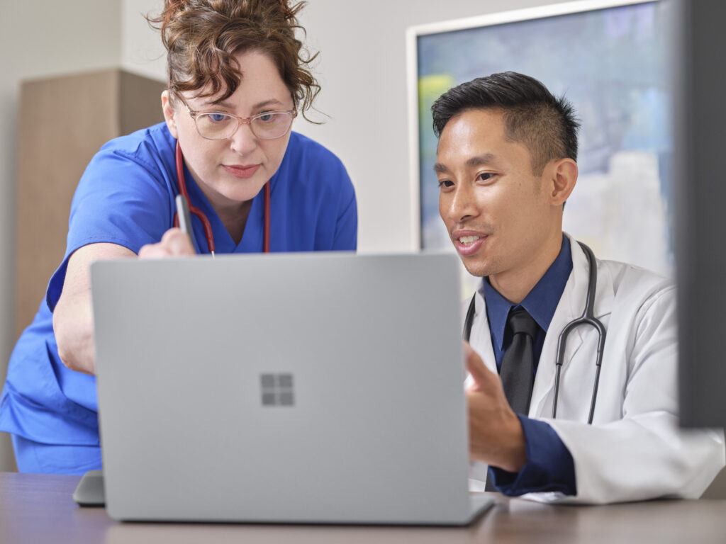 A nurse is pointing out something to the doctor who is working on a Laptop 4.