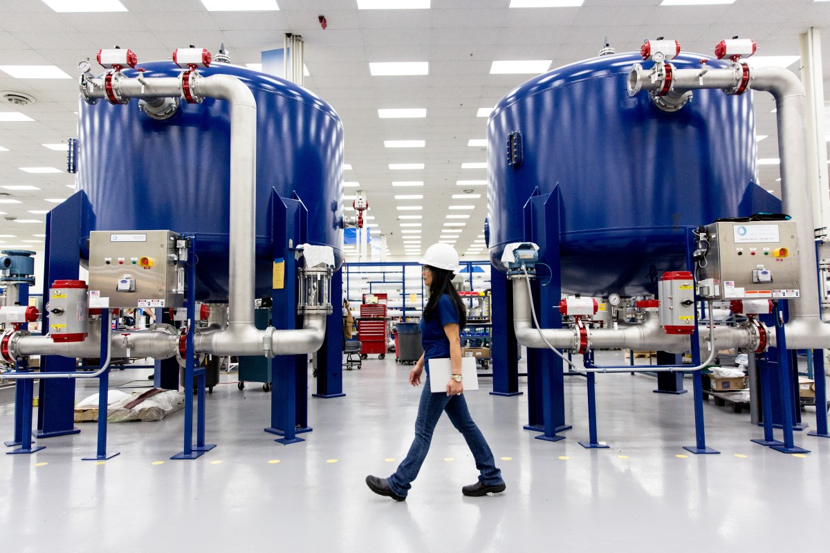 Female worker walking along factory floor in manufacturing plant, in front of two large blue vats. She is wearing a hardhat and safety glasses, and holding a closed tablet by her side