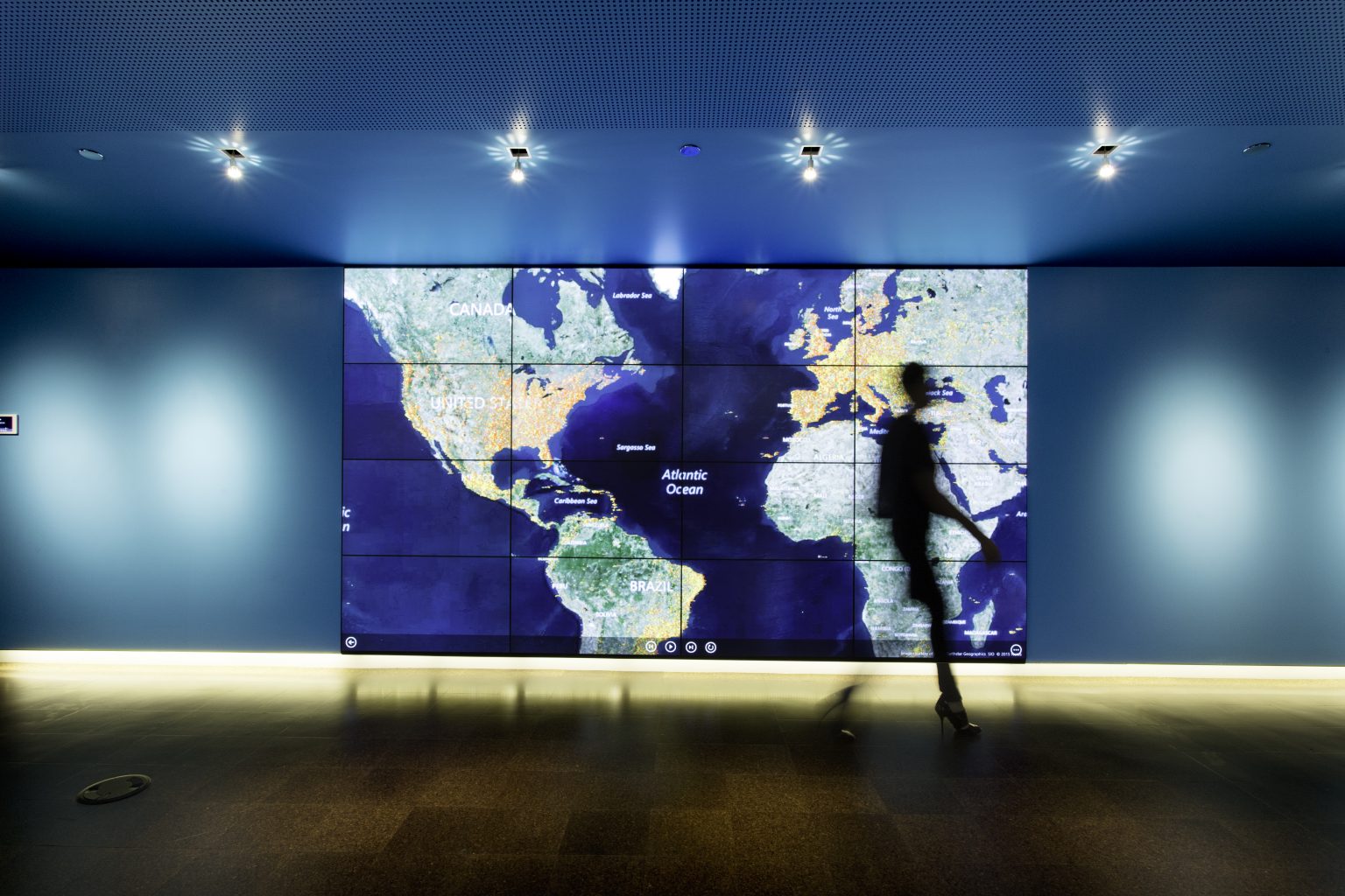 A blurry person walking in front of a large map of the globe on a large screen