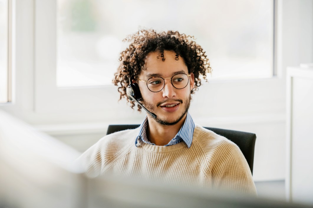 Office employee wearing a wireless headset, talks to clients while working at his computer in the office