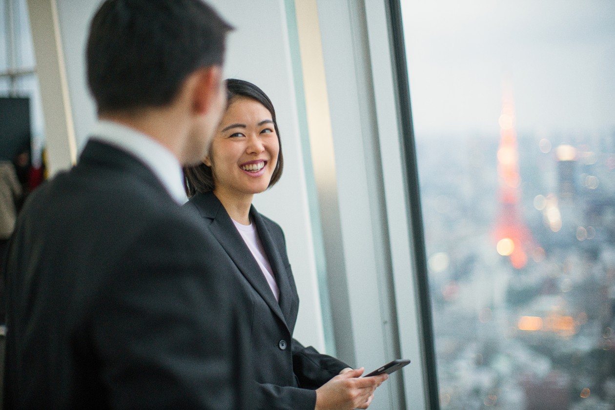 A woman and man, both wearing business attire, stand in front of a window in a tall building. She holds a phone and is smiling