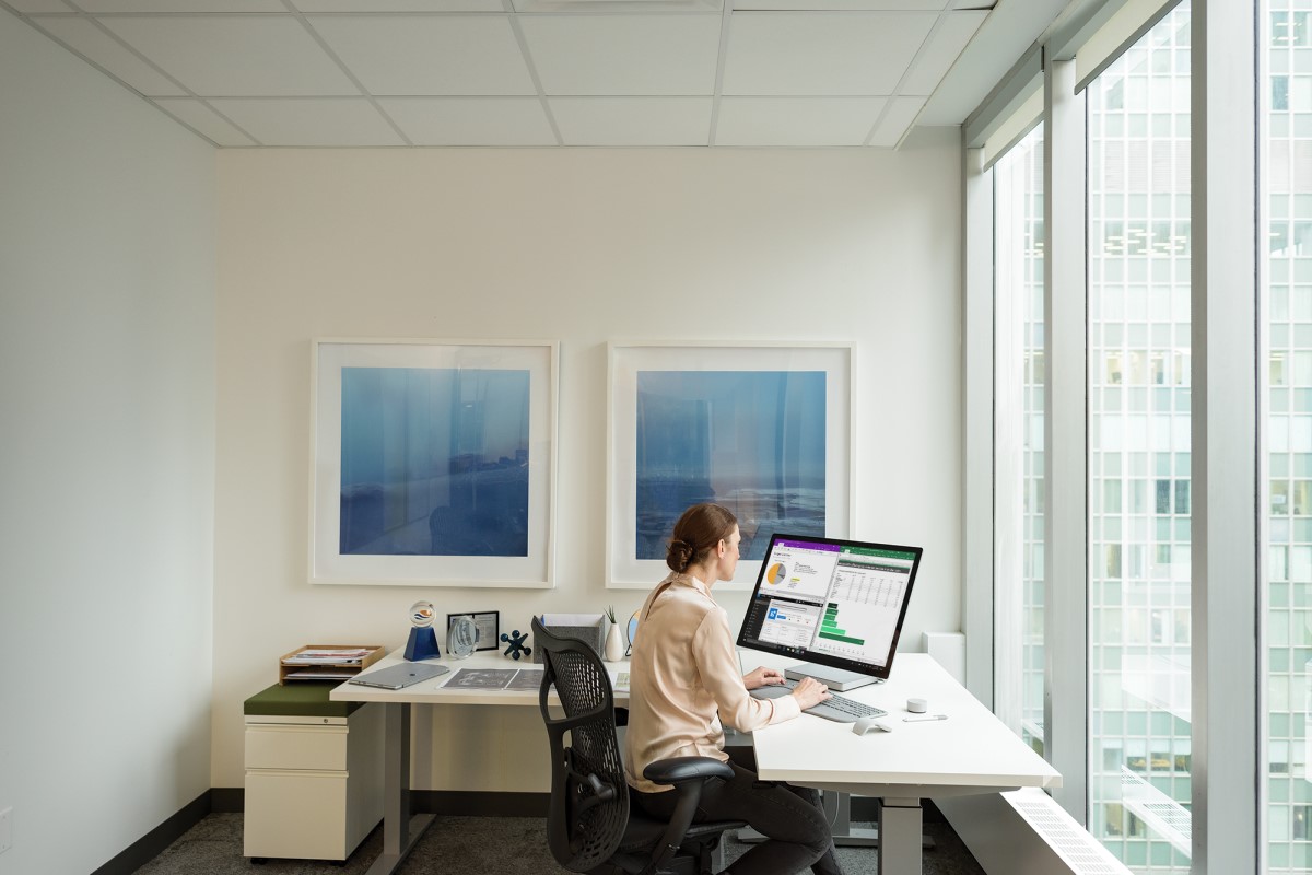 Female office worker seated at corner desk in window office using Surface Studio. Screen shows pie chart and graphs; her hands are on the keyboard