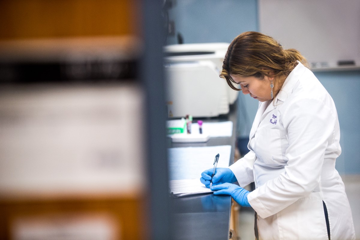 Female technician wearing lab coat and gloves writes with pen and paper in medical laboratory of Chicago hospital