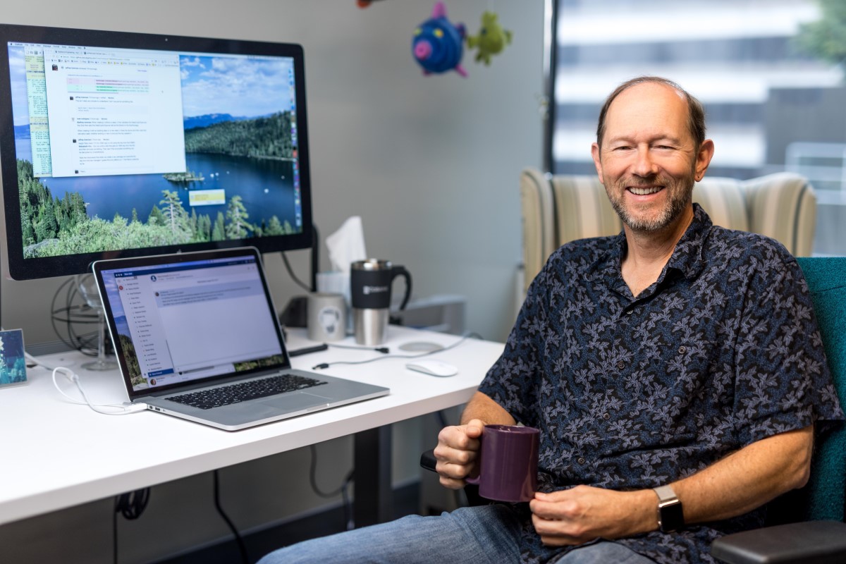 Portrait of older male developer in shirt sleeve shirt, smiling and facing camera. He is sitting at a desk in an office setting, holding a coffee mug