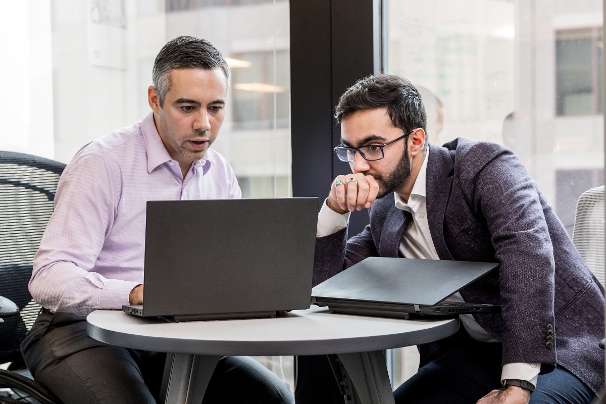 Two male office workers sitting at small round table in financial office, both looking at open laptop (screen not shown)