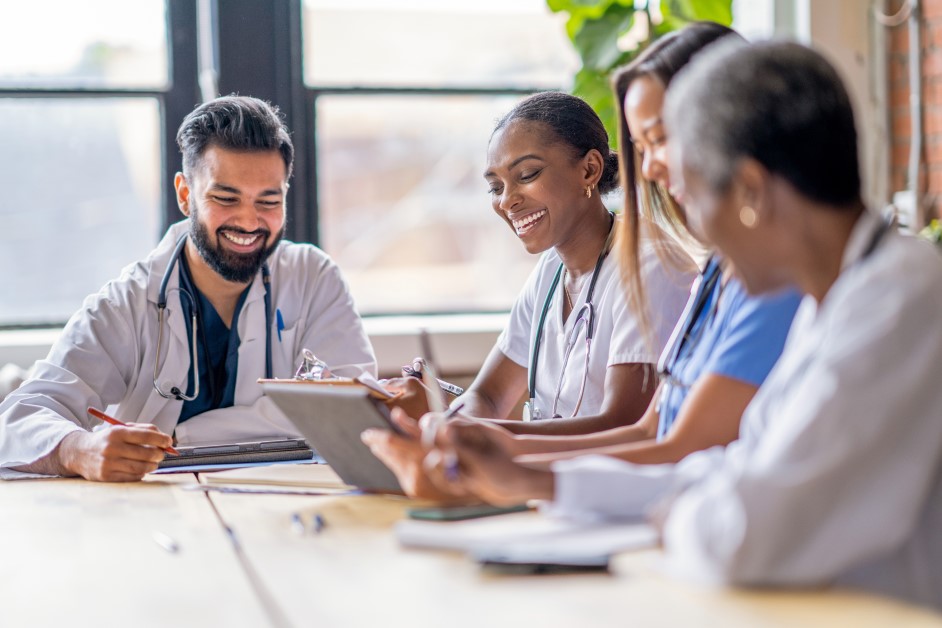 A small group of four medical professionals sit around a conference table as they meet to discuss patient cases. They are each dressed professionally in scrubs and lab coats as they focus on working together
