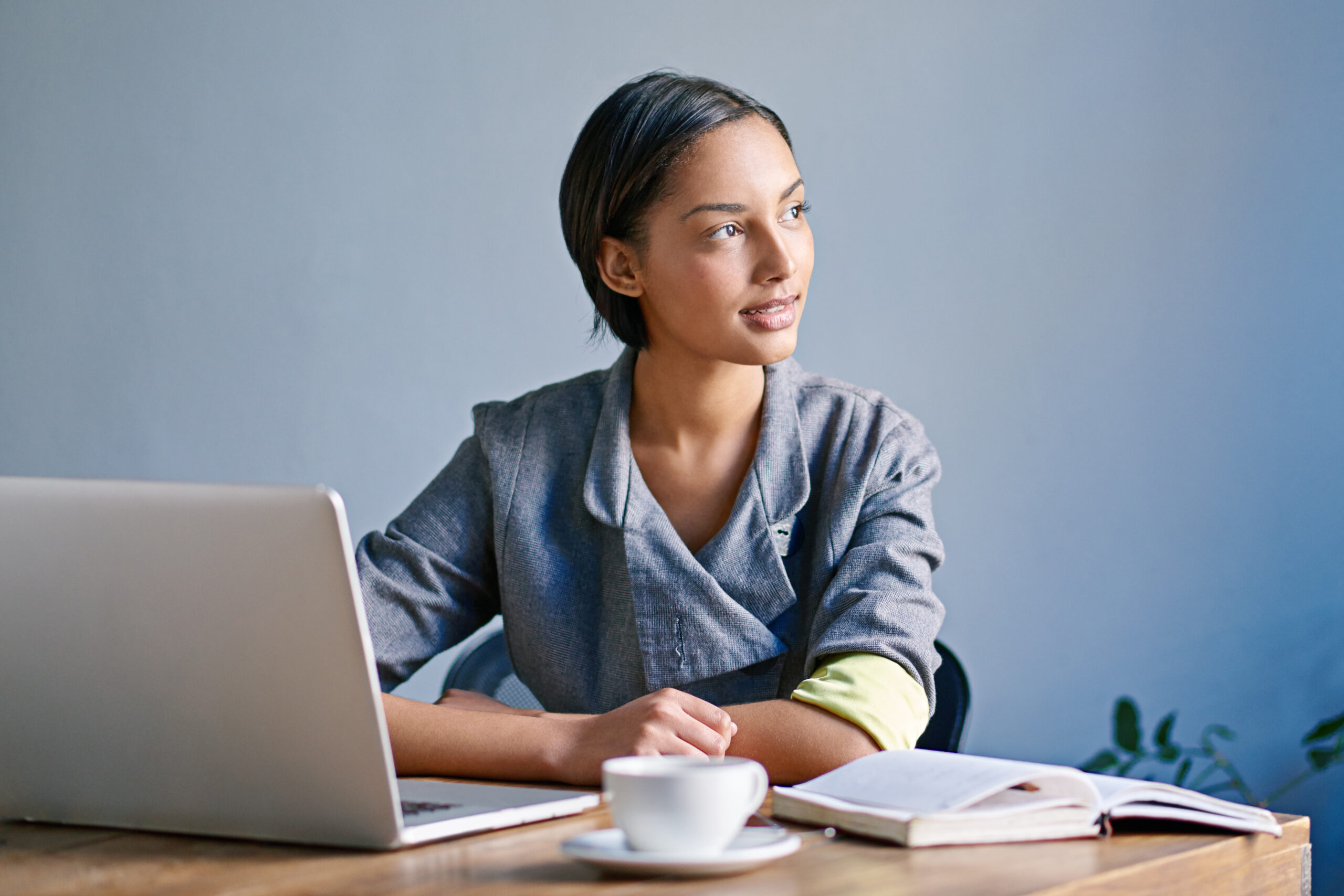 A woman is sitting at a wooden table with a laptop, an open book, and a cup of coffee. The background is plain and the person is wearing a grey jacket