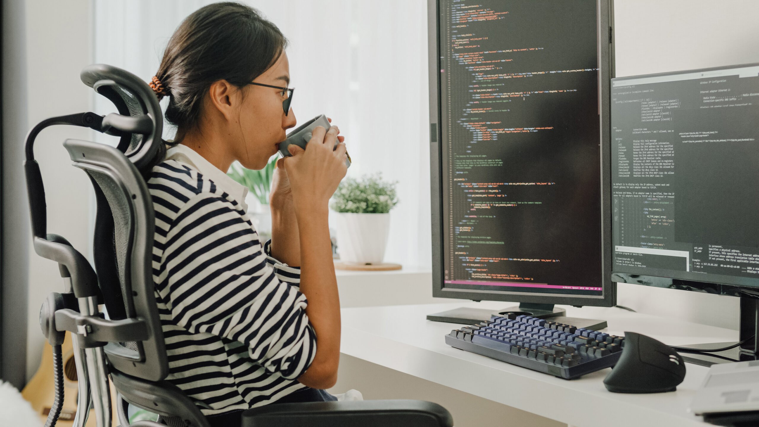 A software developer is using a computer to write code. She is sitting at desk with multiple monitors while drinking coffee