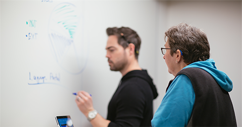 A woman who wears a hearing aid, watches a colleague write on a whiteboard in a conference room