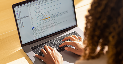 An image of a Black female developer coding on a MacBook Pro in the office, using Visual Studio