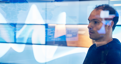 An image of a man in hooded sweater/sweatshirt inside a secure room who is looking at data and a geographic area displayed on a large monitor which is behind glass walls with reflections