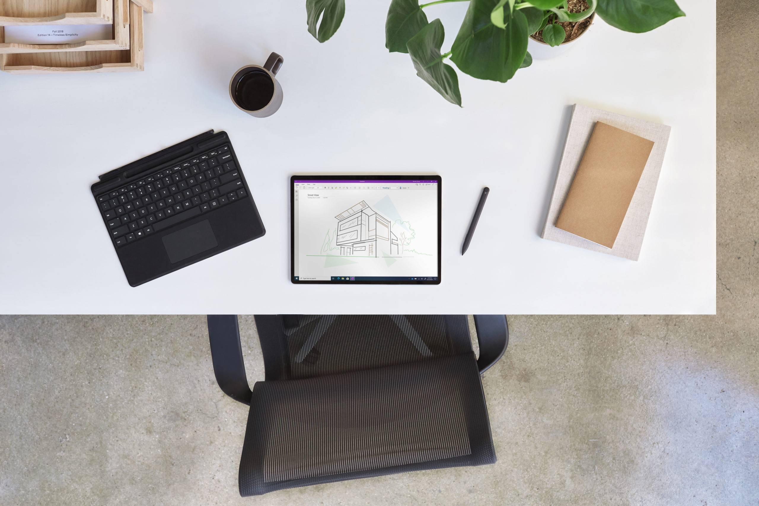 Bird&lsquo;s eye view of Surface Pro X projecting OneNote with a detached black keyboard, pen, books, plant, mug, and wooden shelf