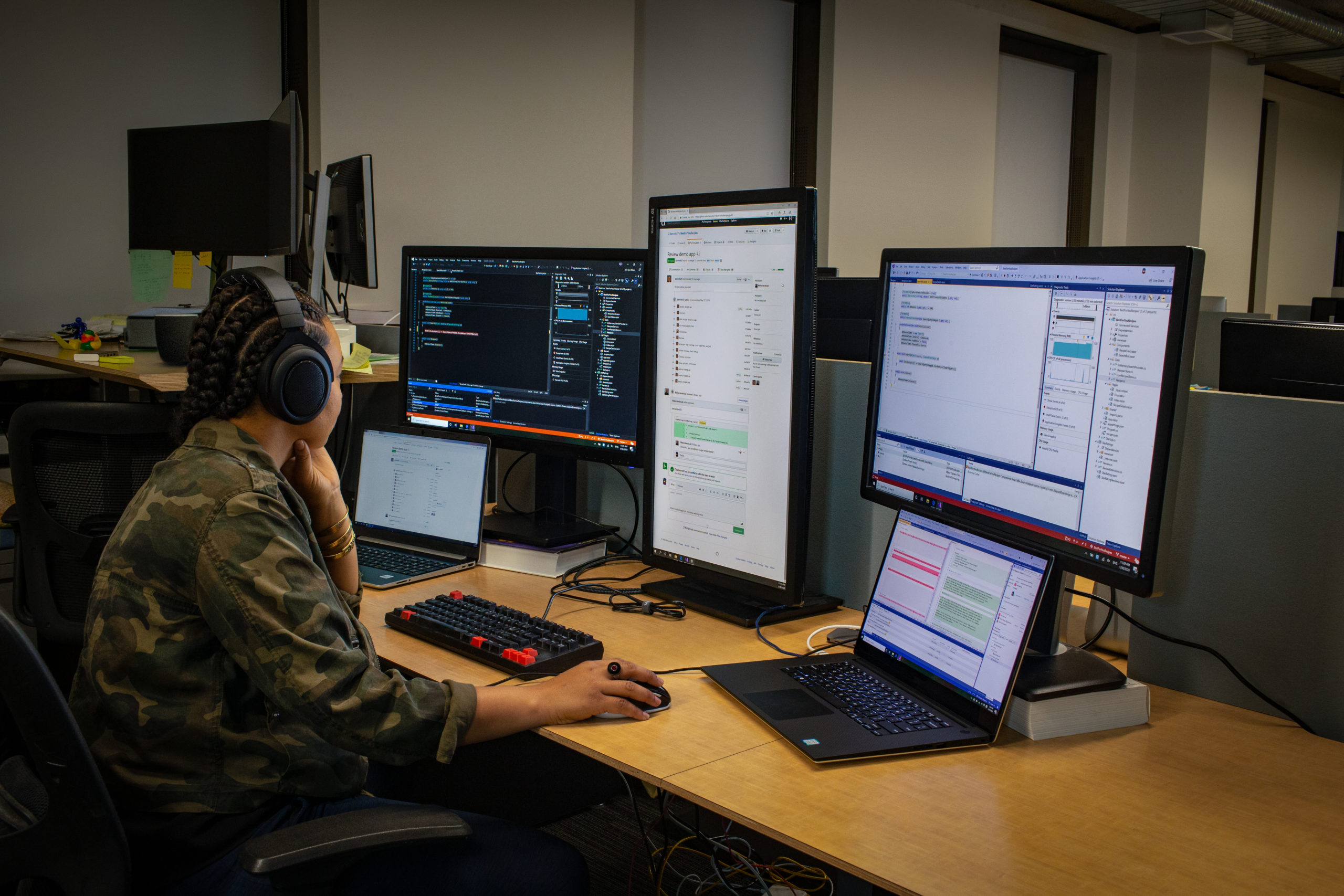 Black female developer wearing headphones; coding at her PC workspace in an enterprise office, using Visual Studio on a multi-monitor set up