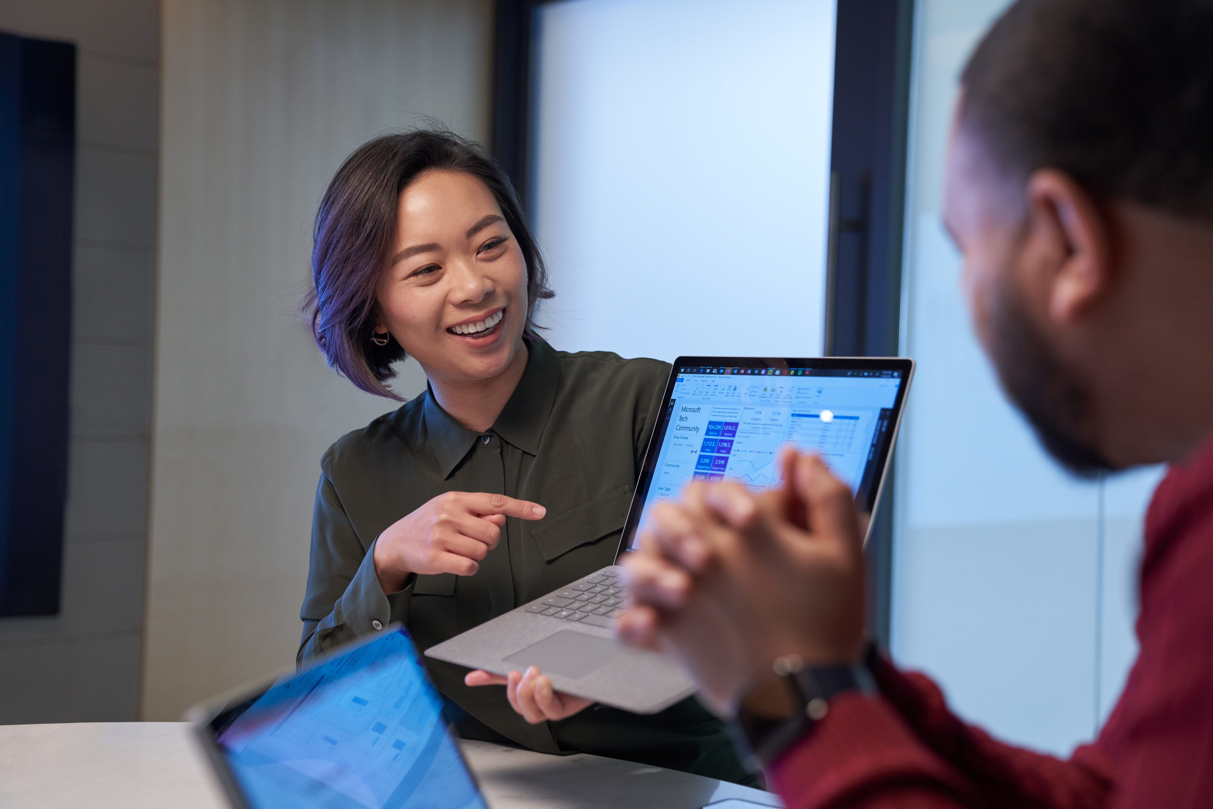 Smiling woman in a meeting room holding up a Surface laptop presenting statistics. Blurred man in foreground