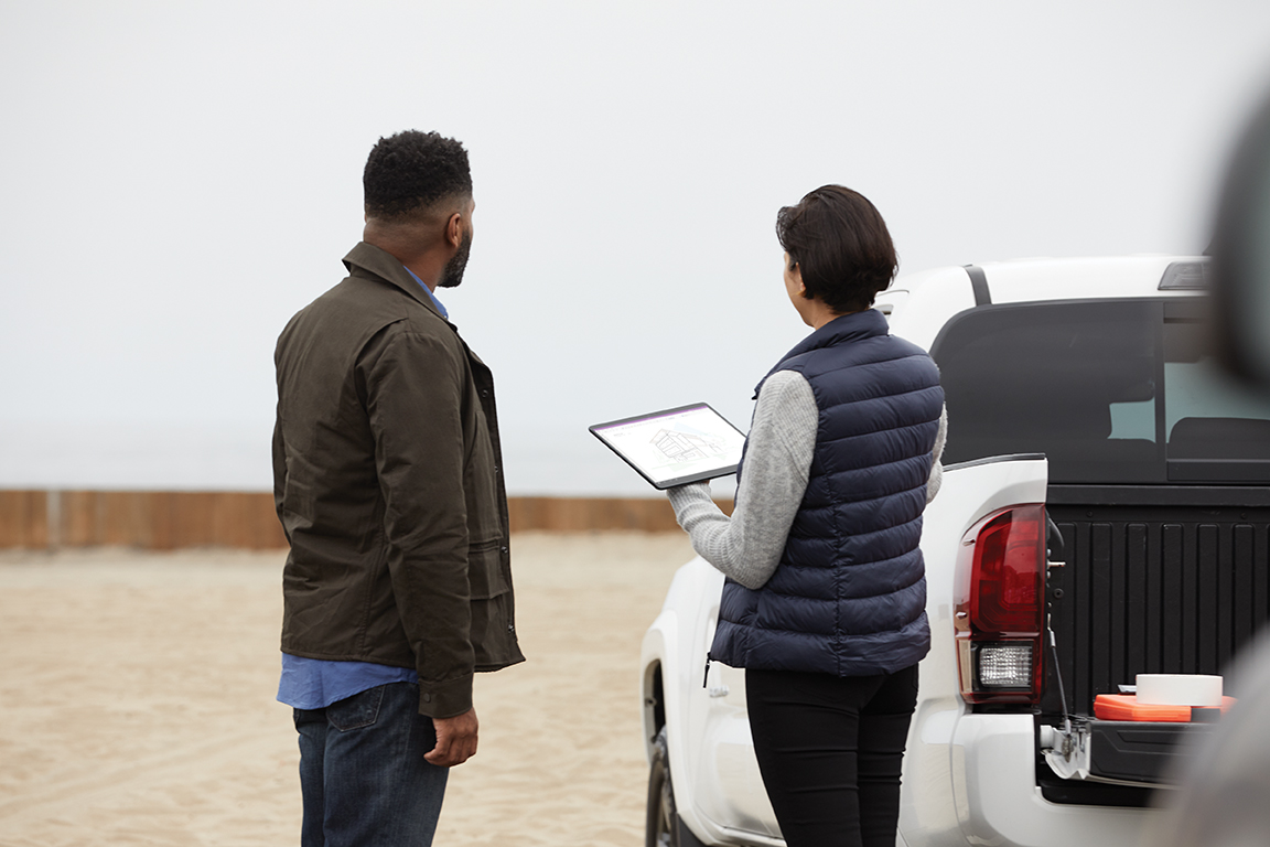 Two adults outside collaborating at a work site while using Microsoft OneNote. Female holding a black Microsoft Surface Pro X in tablet mode with male at her side
