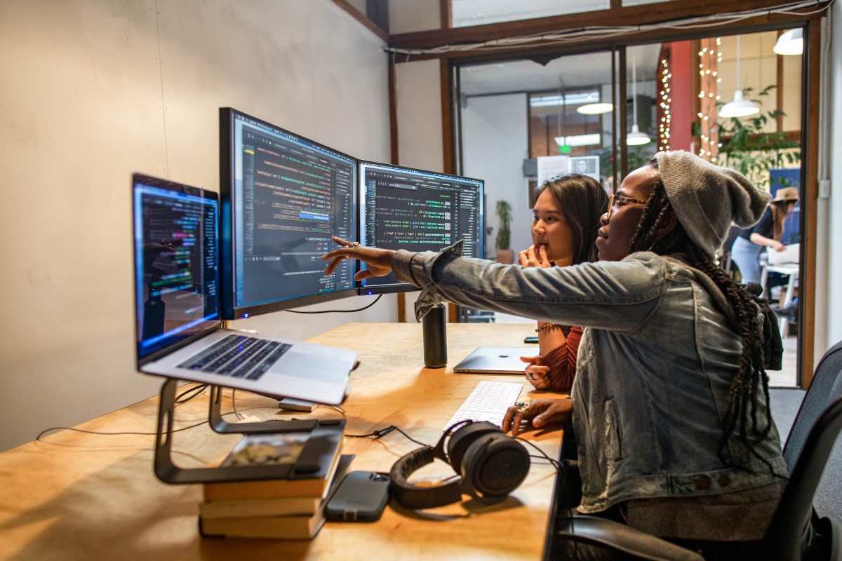 Two female developers working and collaborating in an enterprise office. They are using Visual Studio on a MacBook Pro with a multi-monitor set up