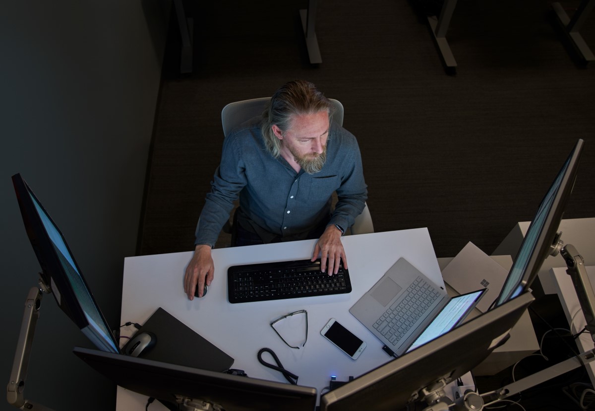 Top-down view of a bearded man in a gray/blue shirt seated at a desk working on a Surface laptop connected to three large monitors