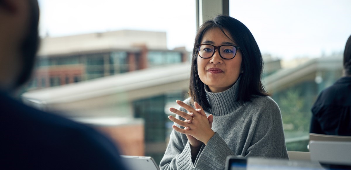 Woman with glasses sitting at a community table in common area looking up from behind a Surface laptop with her hands clasped together