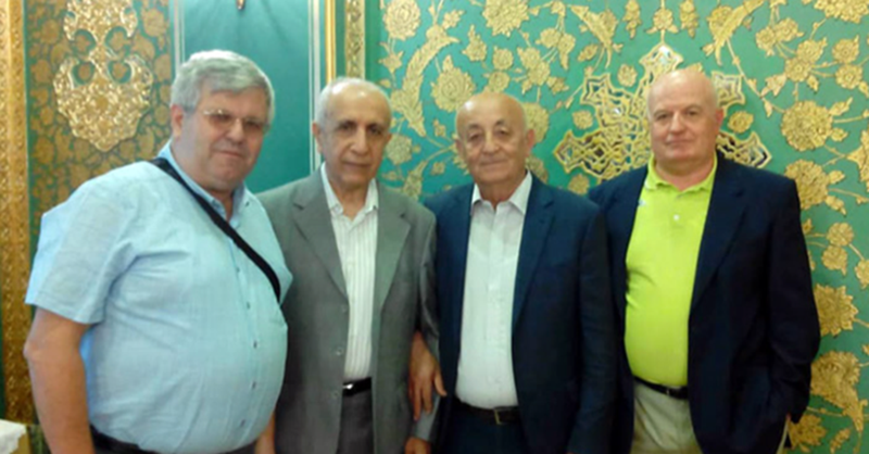 A photo of four males standing closely together, wearing formal clothing and looking at the camera. The male on the leftmost side is wearing glasses.