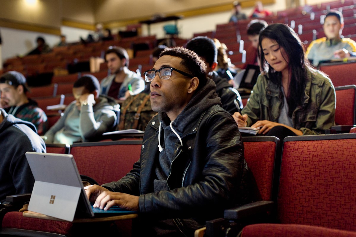 Close up of male college student sitting in university lecture hall, looking ahead while taking notes on a Surface Pro. Other male and female students sit behind him, taking notes