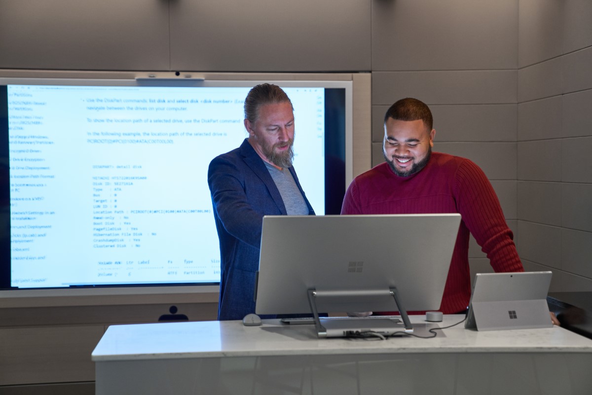 Forward facing view of two men working on a Microsoft Surface Studio with a larger blurred screen and display behind them
