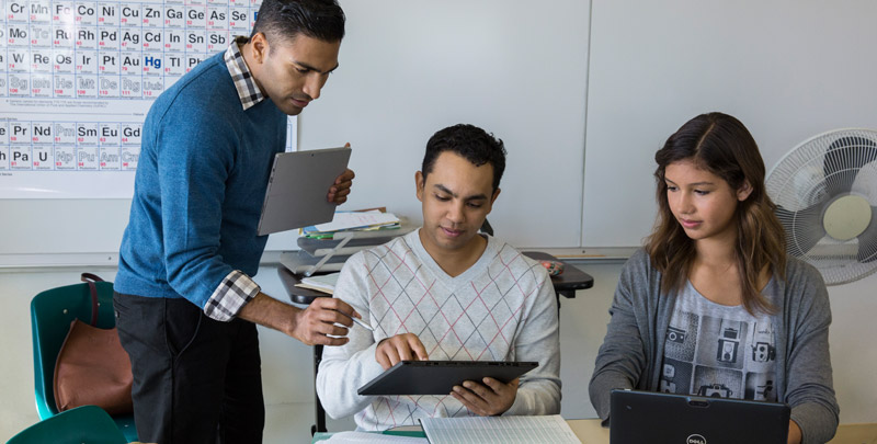Education Color Photo of male educator using device with 6th-9th classroom of male and female students at desks with devices