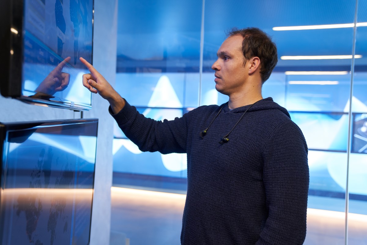 Man in a hooded sweatshirt inside a secure room, pointing at a geographic area displayed on a large monitor