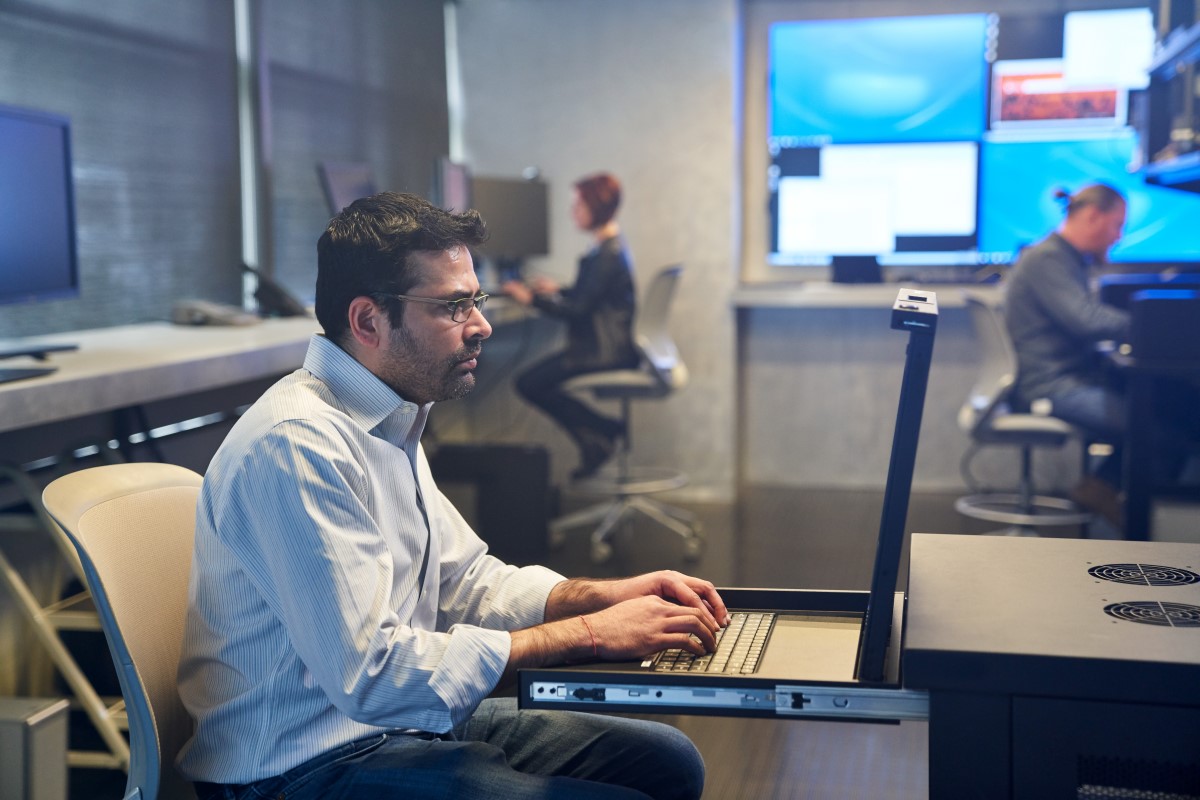 Man in a collared shirt working on a server station inside a secure room. Coworkers and large monitors are in the background