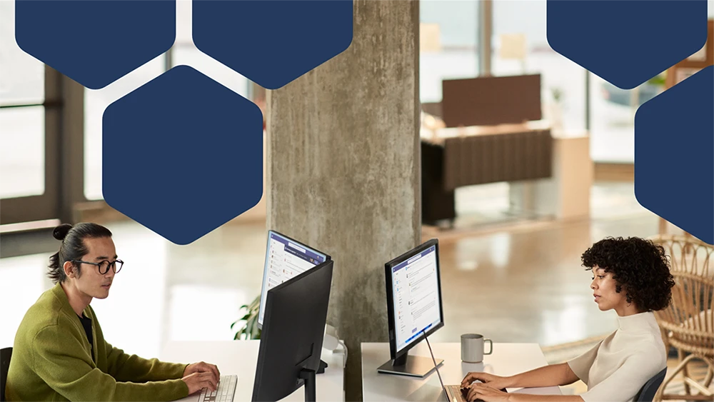 Photo of two employees collaborating during a Microsoft Teams meeting while working in an open office setting on dual monitors