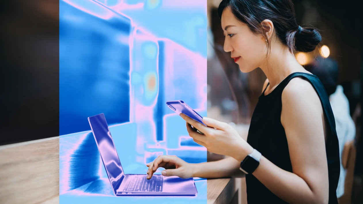 A woman holds a smartphone and types on a laptop