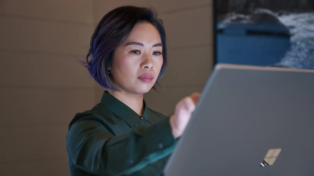 Side profile of a woman wearing a dark shirt in a dim office reaching up and working on a Microsoft Surface Studio