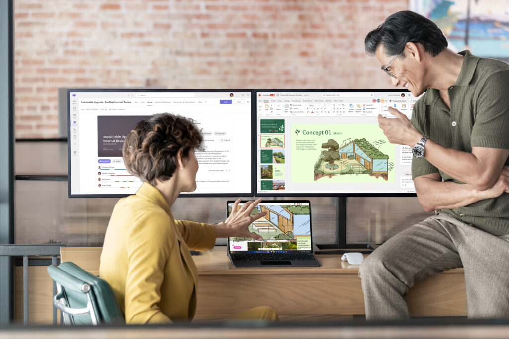 Un homme et une femme collaborent dans un environnement de bureau moderne. La femme, assise, interagit avec un ordinateur portable, tandis que lhomme, debout &agrave; c&ocirc;t&eacute; delle, pointe vers un &eacute;cran. Deux grands &eacute;crans affichent des pr&eacute;sentations sur la durabilit&eacute;, avec des dessins de b&acirc;timents &eacute;cologiques et un document dexamen interne ouvert dans Microsoft Teams. Les deux semblent &eacute;changer des id&eacute;es autour de la conception durable