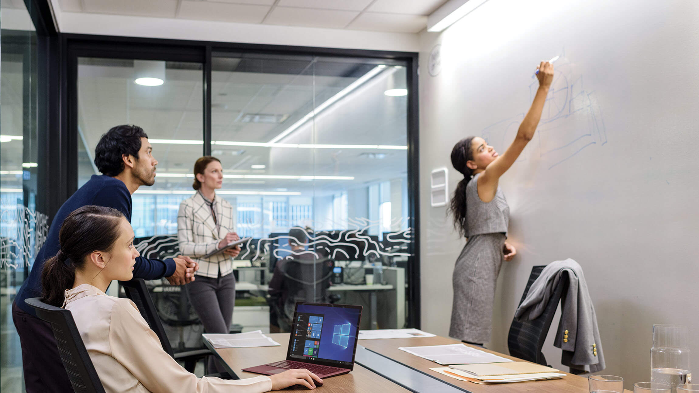 A group of employees in a Microsoft conference room with a person drawing on a white board.