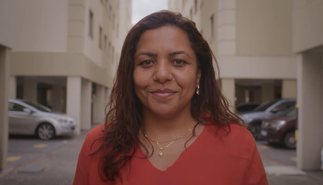 Marcia Pires, Analista de Cloud FinOps, em pé ao ar livre entre prédios de apartamentos, com carros estacionados ao fundo.