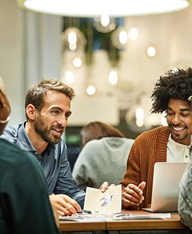 Quatro pessoas conversam ao redor de uma mesa, analisando um laptop em um ambiente com plantas e luzes suspensas.