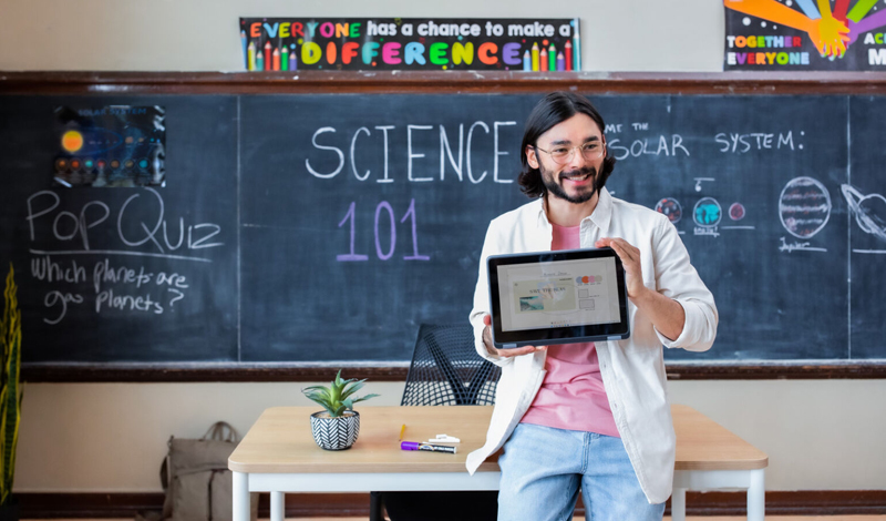 Professor em frente a um quadro negro escrito “Ciências 101”, segurando um tablet com  durante uma aula.